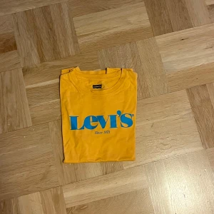 Gul t-shirt från Levi's - Säljer en snygg gul t-shirt från Levi's i storlek 12A. Den har en cool blå logga på framsidan och är gjord i mjuk bomull. Perfekt för sommardagar eller som en färgglad bas i garderoben. 🟡💙