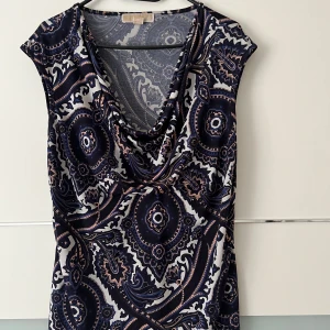 Mönstrad klänning från Michael Kors - Snygg mönstrad klänning från Michael Kors i storlek L. Klänningen har ett unikt mönster i mörkblått och beige med en v-ringad hals. Perfekt för både vardag och festliga tillfällen. Materialet känns mjukt och bekvämt mot huden.