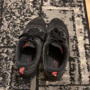 Svarta Nike Air Force sneakers - Säljer ett par svarta Nike Air Force sneakers i bra skick. De har en klassisk design med snörning och en robust sula. Det finns röda detaljer som ger en cool kontrast. Perfekta för vardagsbruk och streetwear-stil.