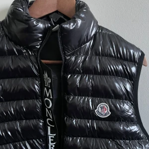 Svart dunväst från Moncler - Säljer en snygg svart dunväst från Moncler i mycket bra skick. Västen har en glansig yta och är quiltad med dragkedja framtill. Perfekt för höst och vår, och den har Monclers logga på bröstet. Passar både till vardags och lite finare tillfällen.