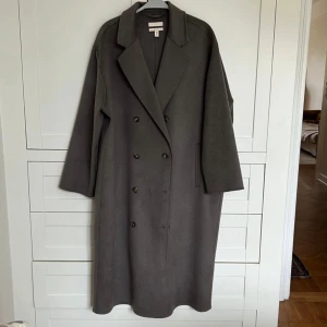 Grå kappa från hm - Säljer en stilren grå kappa från HM i mycket bra skick. Den har en klassisk dubbelknäppt design med långa ärmar och en elegant krage. Storleken är dam M och den är gjord i ett ullmaterial.