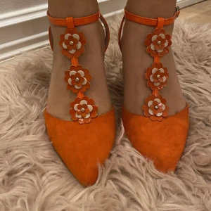 Orange klackskor med blommiga detaljer - Snygga orange klackskor med spetsig tå och blommiga detaljer längs remmarna. Perfekta för en somrig fest eller en utekväll. Skorna har en smal klack och justerbara remmar runt ankeln för bästa passform.