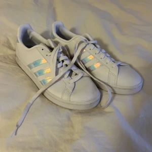 Vita sneakers från Adidas - Säljer ett par vita Adidas sneakers i mycket bra skick. De har de klassiska tre ränderna i en skimrande, nästan holografisk färg. Perfekta för både vardag och fest! Skorna har snörning och är superbekväma. Passar till alla outfits och är ett måste i garderoben. DOM ÄR ALDRIG ANVÄNDA och lappen finns kvar!