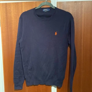 Mörkblå tröja från Ralph Lauren - Säljer en snygg mörkblå tröja från Ralph Lauren i slim fit. Den är gjord av mjuk pima bomull och har en broderad orange logga på bröstet. Bara använd några gånger så den är i väldigt bra skick