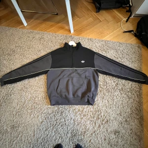 Adidas sweatshirt med dragkedja - Säljer en snygg svart och grå hoodie från Adidas med dragkedja. Den har en klassisk Adidas-logga på bröstet och vita detaljer längs ärmarna. Perfekt för en casual look eller träning. Passar bra till höst och vår.