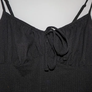 Svart klänning från Gina Tricot - Snygg svart klänning från Gina Tricot i storlek S. Den har smala axelband och en söt knytning framtill. Perfekt för både vardag och festliga tillfällen. Klänningen är i ett mjukt och bekvämt material.