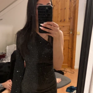 Svart glittrig oneshoulder-klänning - Snygg svart glittrig klänning med oneshoulder-design. Perfekt för festliga tillfällen! Den har en tight passform och långa ärmar med en cool slitsdetalj. Klänningen är både elegant och trendig, idealisk för en utekväll eller nyårsfest.