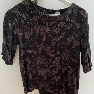 Svart mönstrad topp - Säljer en snygg svart topp med ett unikt mönster i beige. Toppen har trekvartsärmar och en normal passform, perfekt för både vardag och fest. Materialet känns mjukt och bekvämt mot huden. Passar bra till både jeans och kjol!