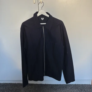 Tunn overshirt/jacka från Arket - Säljer en stilren mörkblå jacka med dragkedja framtill. Perfekt för sommar/vår, med en enkel design som passar till det mesta. Jackan har långa ärmar och en klassisk krage. Den är i bra skick och redo för nya äventyr!