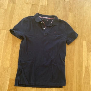 Mörkblå pikétröja från Tommy Hilfiger - Säljer en snygg mörkblå pikétröja från Tommy Hilfiger i storlek 152. Tröjan har korta ärmar och en klassisk krage med knappar. Den är i bra skick och perfekt för både vardag och fest. Den lilla broderade loggan på bröstet ger en stilren touch. Passar perfekt till jeans eller chinos!