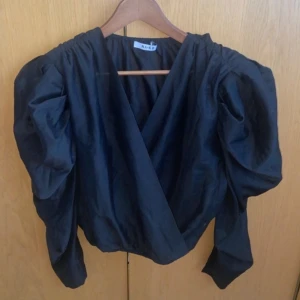 Svart omlottopp från NA-KD - Snygg svart omlottopp från NA-KD med puffiga långa ärmar. Toppen har en elegant omlottdesign som ger en smickrande passform.