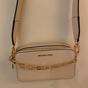 Beige axelväska från Michael Kors - Säljer en superfin beige axelväska från Michael Kors i skinn. Väskan har en elegant kedjedetalj i guld och en bred axelrem med nitar. Perfekt för både vardag och fest! Den har en dragkedja upptill och flera fack inuti för bra organisering. Väskan är i mycket bra skick och redo för nya äventyr. (NYPRIS 2700kr)