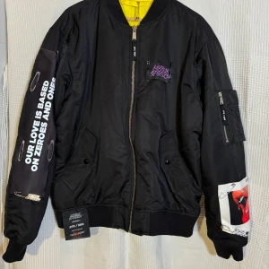 Svart bomberjacka från Marius Sperlich - Säljer en unik svart bomberjacka från Marius Sperlich, nummer 275 av 500. Jackan har en cool design med säkerhetsnålar och ett stort tryck av röda läppar på ryggen. Den har dragkedja fram och flera fickor. Insidan är klargul och materialet är en mix av polyester och återvunnen polyester. 