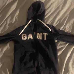 Mörkblå hoodie från GANT - Säljer en snygg mörkblå hoodie från GANT i storlek XS. Den har en dragkedja framtill och stora bokstäver på bröstet. Perfekt för höst och vinter, och den är superbekväm med sin mjuka insida. Passar både till vardags och chillkvällar. Ny pris runt 1000 kr men mitt pris är endast 299 !!