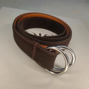 Brunt mockabälte från Polo Ralph Lauren - Exklusivt unisexbälte från Polo Ralph Lauren i mocka – helt nytt!  Säljer ett lyxigt unisexbälte från Polo Ralph Lauren, aldrig använt och i perfekt skick. Perfekt för den som uppskattar hög kvalitet och elegant design. 	•	Material: Brun mocka med läderfoder. 	•	Spänne: Dubbla silverringar. 	•	Storlek: 100 cm (inklusive spänne). 	•	Bredd: 3 cm.  Ett tidlöst och stilfullt tillbehör som passar både kvinnor och män, perfekt både till vardags och fest 