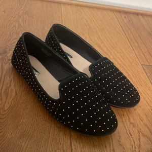 Svarta loafers med prickigt mönster - Säljer ett par snygga svarta loafers med vita prickar. De är perfekta för både vardag och fest. Skorna har en klassisk form och är gjorda i ett mjukt material som ger en bekväm passform.