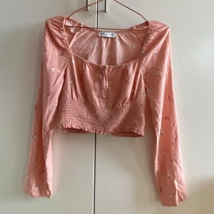 Rosa blommig topp med smockdetalj - Säljer en superfin rosa topp med blommönster och smockdetalj i midjan. Toppen har långa ärmar och en söt knytning framtill. Perfekt för vår och sommar!