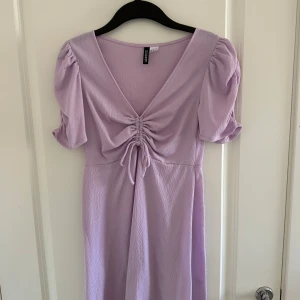 Ljuslila klänning från H&M Divided - Säljer en superfin ljuslila klänning från H&M Divided. Den har en v-ringad design med snygg snörning framtill och korta puffärmar. Materialet är mjukt och bekvämt, perfekt för både vardag och fest. Klänningen är i nyskick och redo för nya äventyr!