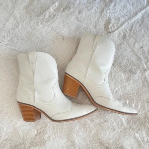 Vita cowboy boots med klack - Säljer ett par snygga vita låga cowboy boots i skinn med en träfärgad klack. De har en spetsig tå och dekorativa sömmar som ger en cool look. Perfekta för både vardag och fest!