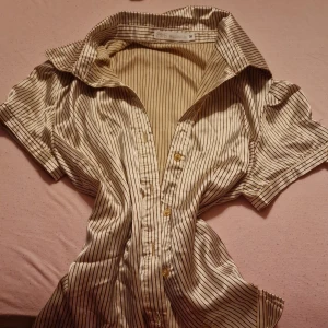 Beige och svart randig skjorta från Millenium - Säljer en snygg beige skjorta med svarta ränder från Millenium. Skjortan har korta ärmar och knappar framtill. Perfekt för både vardag och fest! 🖤✨