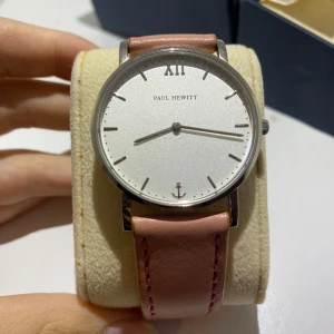 Elegant klocka från Paul Hewitt - Säljer en stilren klocka från Paul Hewitt med en klassisk vit urtavla och romerska siffror. Klockan har en snygg rosa läderrem med rosa sömmar, vilket ger den en modern touch. Tillverkad i Tyskland och en del av deras Sailor Line. Perfekt för både vardag och fest!