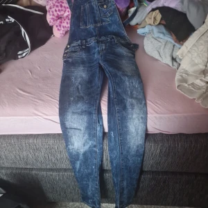 Blå jeansbyxdress - Säljer en snygg blå jeansbyxdress med hängslen och flera fickor fram. Perfekt för en avslappnad stil och kan matchas med en t-shirt eller tröja under. Byxdressen har en klassisk jeanslook med slitna detaljer.