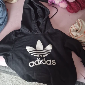 Svart hoodie från Adidas - Säljer en svart hoodie från Adidas med den klassiska loggan i vitt på framsidan. Den har en stor och bekväm huva samt en praktisk magficka. Perfekt för en avslappnad stil eller träning. Hör av dig om du är intresserad!