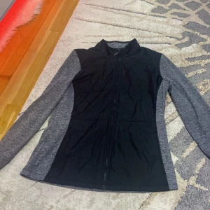 Grå och svart jacka från Cutter & Buck - Säljer en snygg och funktionell jacka från Cutter & Buck i storlek L. Den är grå med svarta detaljer och har en dragkedja framtill. Perfekt för höst och vår, med långärmade ärmar och en stilren design. Materialet känns bekvämt och passar bra för både vardag och träning.