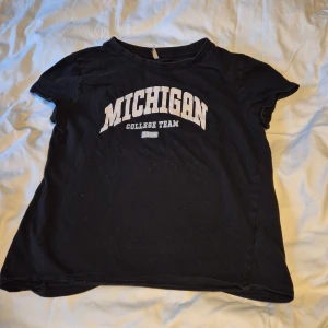 Svart t-shirt med Michigan-tryck - Säljer en svart t-shirt med trycket 'Michigan College Team' i vitt. Den är i storlek 158/164 och passar perfekt för en avslappnad stil. T-shirten är kortärmad och gjord i ett mjukt bomullsmaterial. Perfekt för vardagsbruk eller sportiga tillfällen!