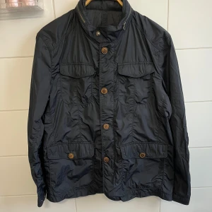 Marinblå jacka från Massimo Dutti - Säljer en snygg marinblå jacka från Massimo Dutti i storlek XL. Jackan har en stilren design med knappar och dragkedja. Den har även en inbyggd luva som man kan veckla ut och in när man vill. Frågor och funderingar? Hör bara av dig.
