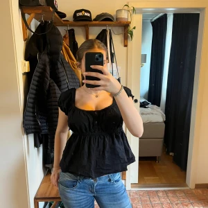 Svart blus med knappar - Säljer en snygg svart blus med knappar framtill. Den har en luftig passform och korta ärmar, perfekt för både vardag och fest. Blusen är i bra skick och passar till jeans eller kjol. Perfekt för en stilren look!
