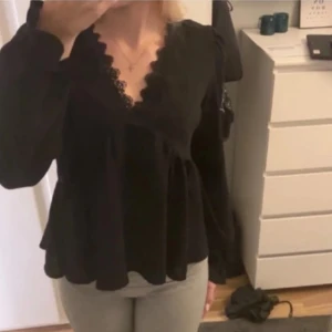 Svart blus med spetsdetaljer - Säljer en elegant svart blus med v-ringad spetsdetalj. Blusen har långa ärmar och en volangkant nedtill som ger en fin siluett. Perfekt för både vardag och fest!