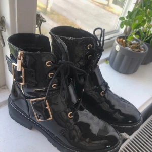 Svarta lackade boots med spänne och snörning - Säljer ett par snygga svarta boots i lackat material. Perfekta för höst och vinter när du vill ha något som sticker ut! De är i nyskick knappt använda har förvarats i garderoben och går ej att köpa någon stans längre så de är väldigt unika. 