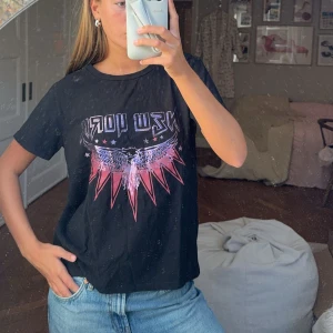 Svart t-shirt med New York-tryck - Säljer en svart t-shirt med coolt New York-tryck och en färgglad örn i rosa och lila. Perfekt för en avslappnad stil. T-shirten är kortärmad och i bomull, vilket gör den skön att bära till vardags. Passar perfekt till jeans för en casual look.