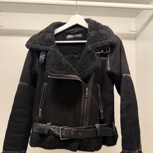 Svart jacka från Zara - Säljer en supercool svart jacka från Zara i storlek XS. Den har en snygg bikerstil med dragkedja och bälte i midjan. Jackan är fodrad med mysig teddy och har långa ärmar. Perfekt för höst och vinter när du vill hålla dig varm och stilren. 🖤