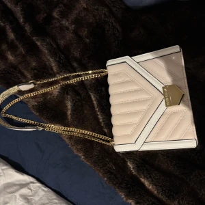 Beige quiltad axelväska med guldkedja - Säljer en superfin beige axelväska med quiltad design och guldkedja. Väskan har en snygg geometrisk detalj framtill och är perfekt för både vardag och fest. Den är i nyskick och redo att användas direkt!