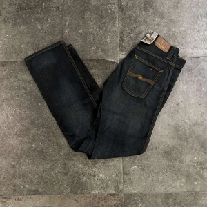 Mörkblå jeans från Nudie Jeans - Helt nya snygga mörkblå jeans från Nudie Jeans i slim/straight fit. Tillverkade av 100% ekologisk bomull med klassiska femficksdetaljer och kontrastsömmar. Perfekta för en stilren look. Jeansen har lappen kvar och är storlek 30/34, nypris ca 1700kr dock så görs inte dessa längre, mitt pris 599kr. fraktas antigen samma dag som köp eller dagen därpå💯