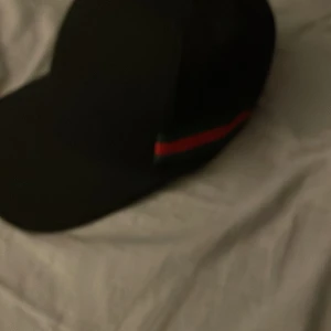 Svart keps med ränder - Snygg svart keps med gröna och röda ränder på sidan. Kepsen har en justerbar rem baktill för perfekt passform. Perfekt för en stilren look.