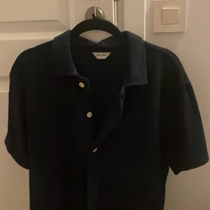 Mörkblå kortärmad sommar skjorta - Säljer en stilren mörkblå pikétröja från East West. Tröjan har korta ärmar och knappar framtill. Perfekt till sommaren, tröjan har även riktigt skönt och mjukt material. Nypris 700 mitt pris 280 men pris kan även diskuteras!