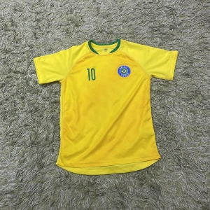 Gul Brasilien fotbollströja från H&M - Säljer en gul Brasilien fotbollströja från H&M med gröna detaljer och nummer 10 på bröstet. Tröjan har korta ärmar och ett runt halsmärke med texten 'Brasil'. Perfekt för fotbollsfans! 💛💚