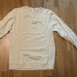 Vit sweatshirt från Les Deux - Säljer en stilren vit sweatshirt från Les Deux med broderad logga på bröstet. Tröjan har långa ärmar och en klassisk rund halsringning. Perfekt för en avslappnad stil.