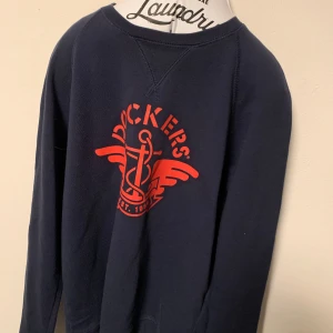 Mörkblå sweatshirt från Dockers - Säljer en mörkblå sweatshirt från Dockers med ett stort rött tryck framtill. Tröjan har långa ärmar och rund halsringning. Perfekt för en avslappnad stil. Storlek M.