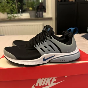 Nike Presto - Snygga Nike Presto sneakers i svart med blå detaljer. Skorna har en modern design med snörning och en bekväm passform. Perfekta för en stilren look.