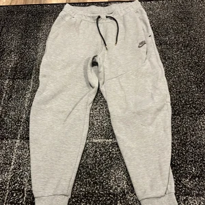 Grå mjukisbyxor från Nike - Snygga grå mjukisbyxor från Nike med elastisk midja och snörning. Byxorna har en liten Nike-logga på sidan och är perfekta för en avslappnad stil. De har muddar vid bensluten för en bekväm passform.