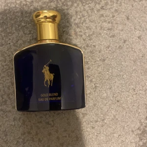 Gold Blend Eau de Parfum - Elegant och lyxig parfym i en djupblå flaska med gyllene detaljer. Flaskan har en ikonisk logotyp med en polospelare och en gyllene kork. Perfekt för den som vill ha en sofistikerad doftupplevelse.