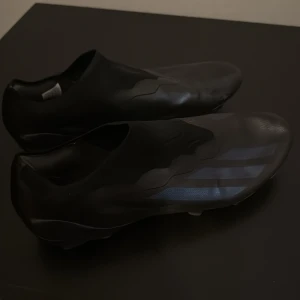 Svarta fotbollsskor med blå detaljer - Snygga svarta fotbollsskor med diskreta blå detaljer. Skorna har en strömlinjeformad design utan snörning för en tight passform. Perfekta för spel på gräsplaner. Använda kanske 10 gånger litet skrap på framsida på skorna men inget farligt. Frakt kan även åka själv och lämna 