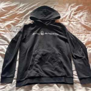 Svart hoodie från Sail Racing - Säljer en svart hoodie från Sail Racing med vit logga på bröstet. Den har en klassisk känguruficka och en bekväm huva. Perfekt för en avslappnad stil.
