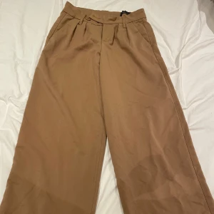 Bruna kostymbyxor med knappar - Snygga bruna kostymbyxor med låg midja och vida ben. Byxorna har knappar och dragkedja framtill samt hällor för bälte. Perfekta för en stilren look.💕