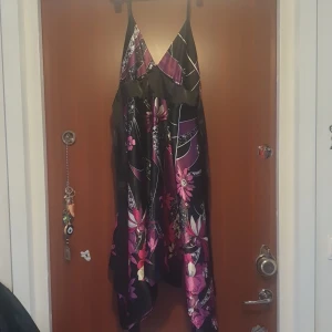 Svart och lila blommig klänning - Snygg svart klänning med lila och rosa blommönster. Den har en v-ringad design med smala axelband och ett band i midjan. Perfekt för en färgglad look!