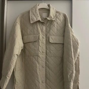 Beige  jacka från H&M - Säljer en stilren beige quiltad jacka från H&M. Jackan har långa ärmar och knappar framtill samt två stora fickor på bröstet. Perfekt för kyligare dagar och ger en trendig look.